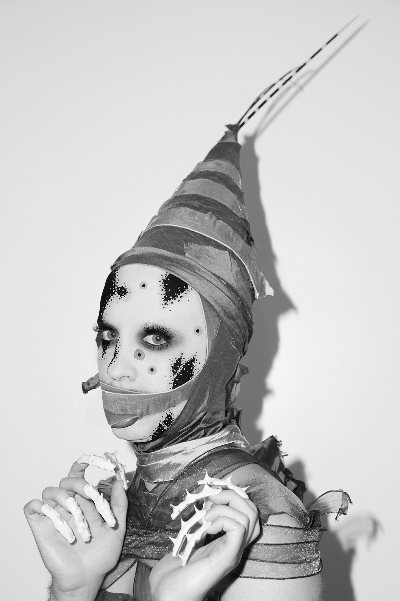 Jesse with graphic black and white facepaint on, his mouth covered with a strip of farbic and a fabric cone on his head against a white background